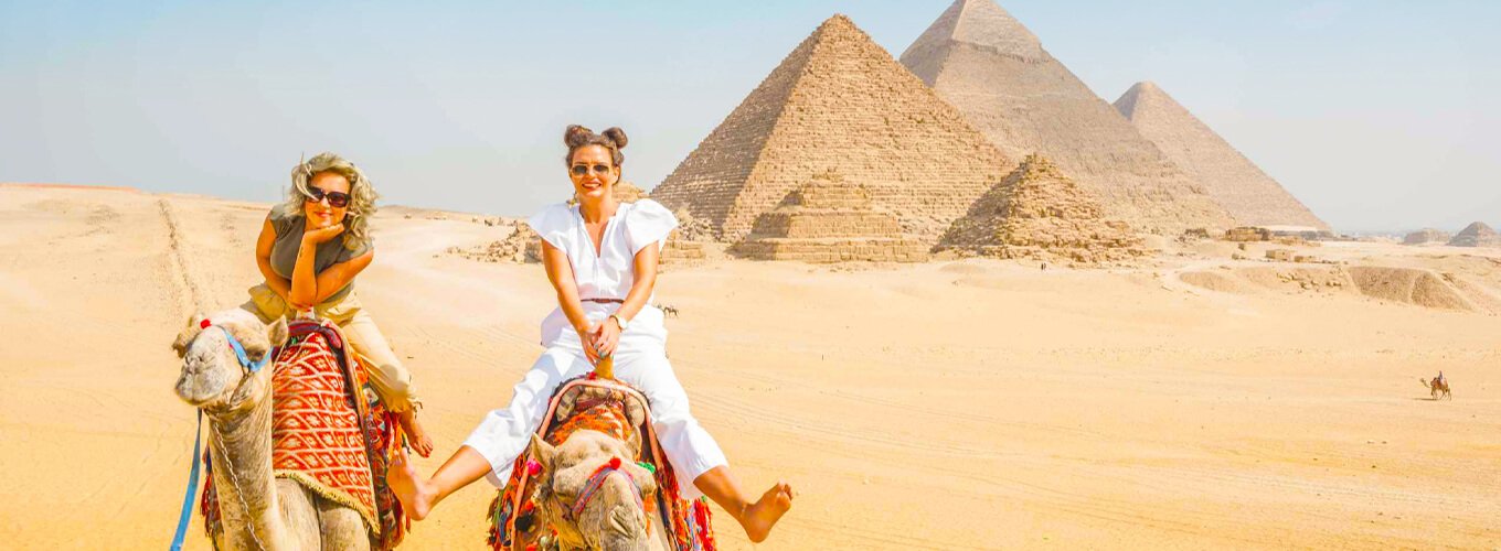 Dos niñas montando un camello frente a las pirámides de Egipto.